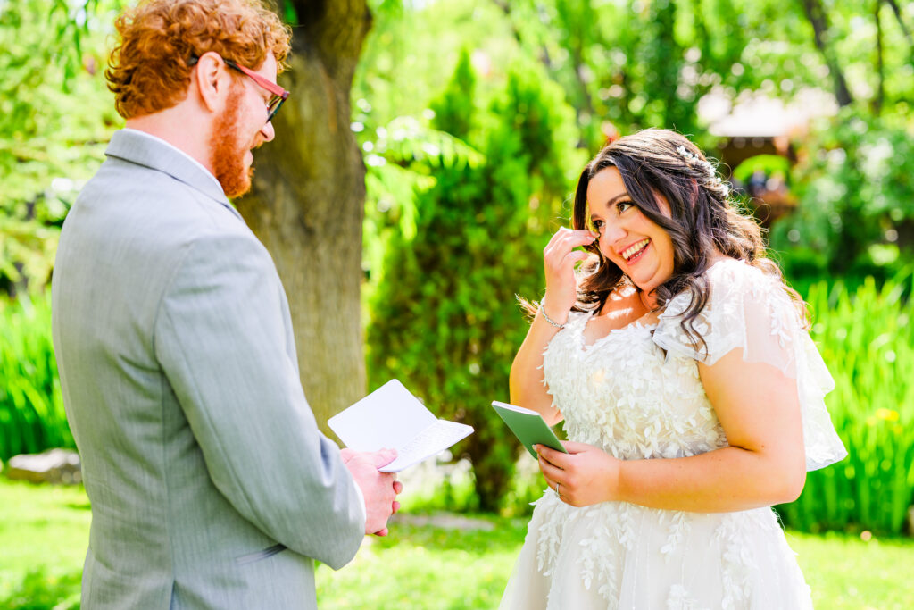 bride crying wiping her tears as groom reads vows during private moment between bride and groom with green forest background on lawn at The Windmill House in Chino Valley Prescott Arizona 